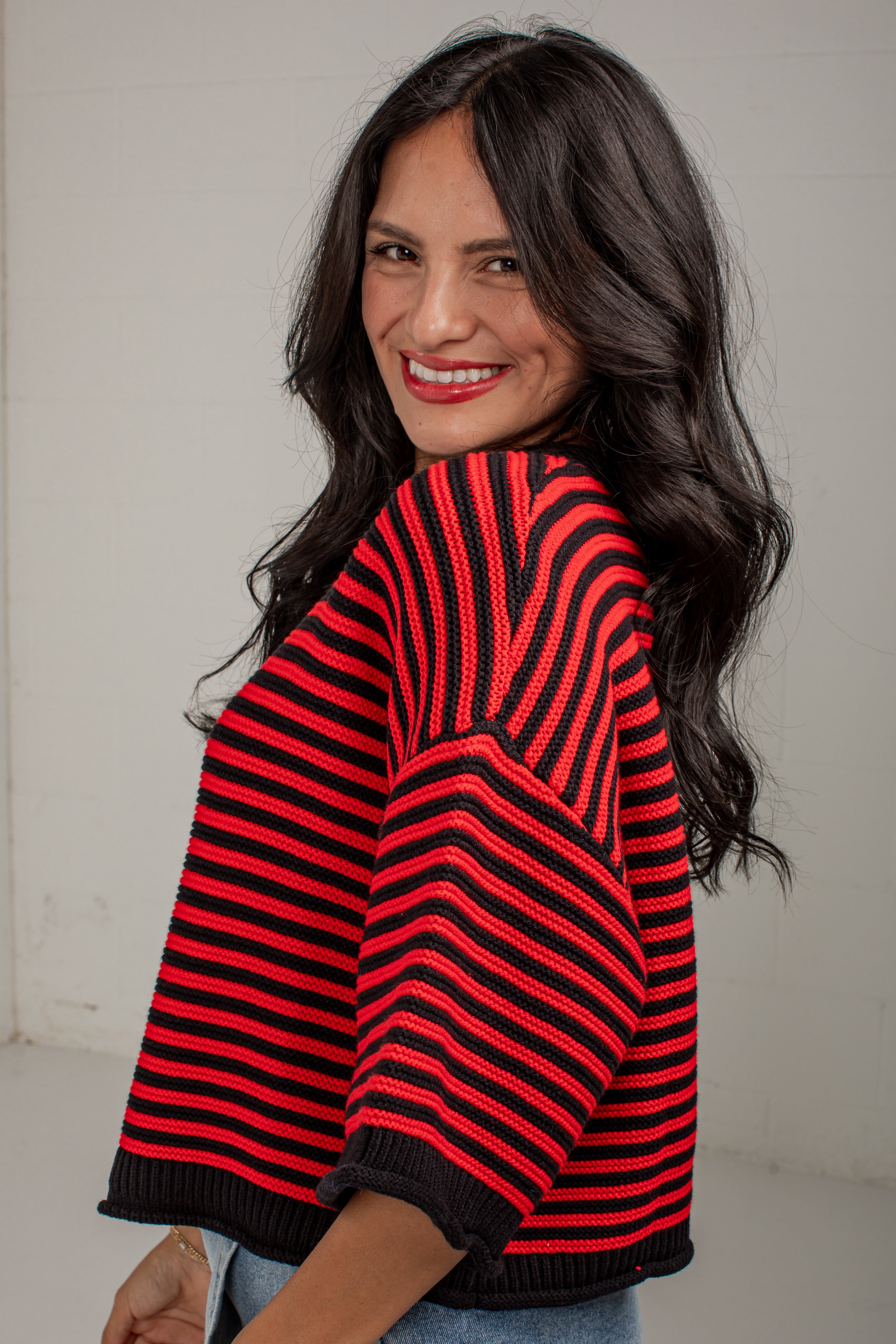 A woman with long dark hair is shown from the back and side, wearing a red and black striped knit sweater with wide three-quarter sleeves and a ribbed hem, against a plain light gray wall.