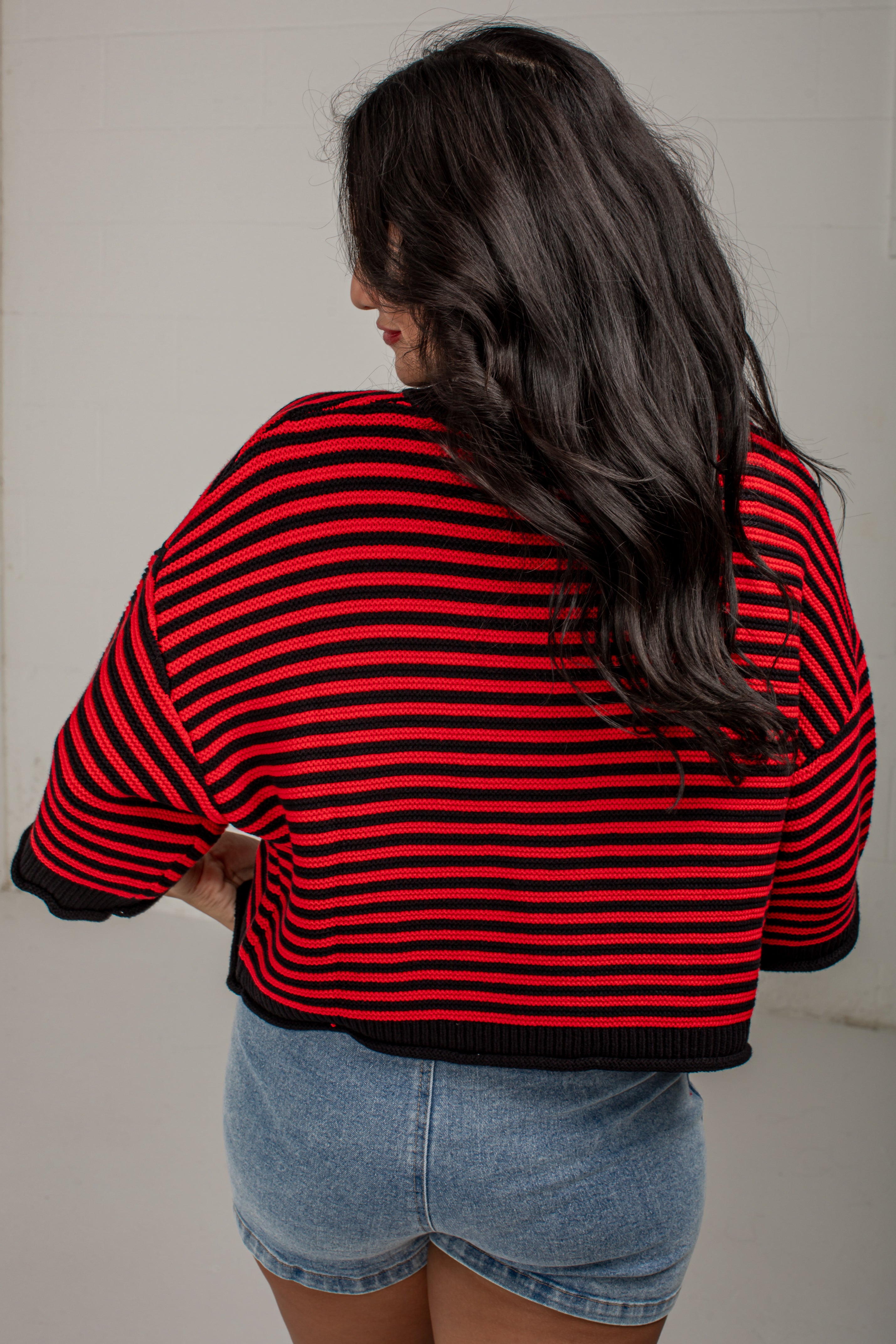 A woman viewed from behind wearing a red and black horizontally striped cropped knit sweater with black trim, paired with light blue denim shorts, against a plain off-white wall.