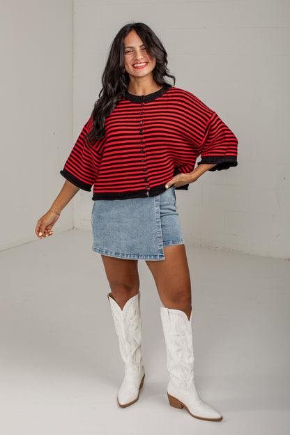 A woman modeling a red and black striped knit poncho-style cardigan with a button front, paired with a light blue denim skirt and white cowboy boots, in a bright studio setting.