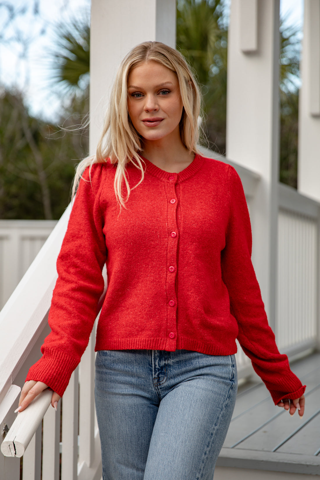 Woman wearing a red cardigan and blue jeans standing on a porch.