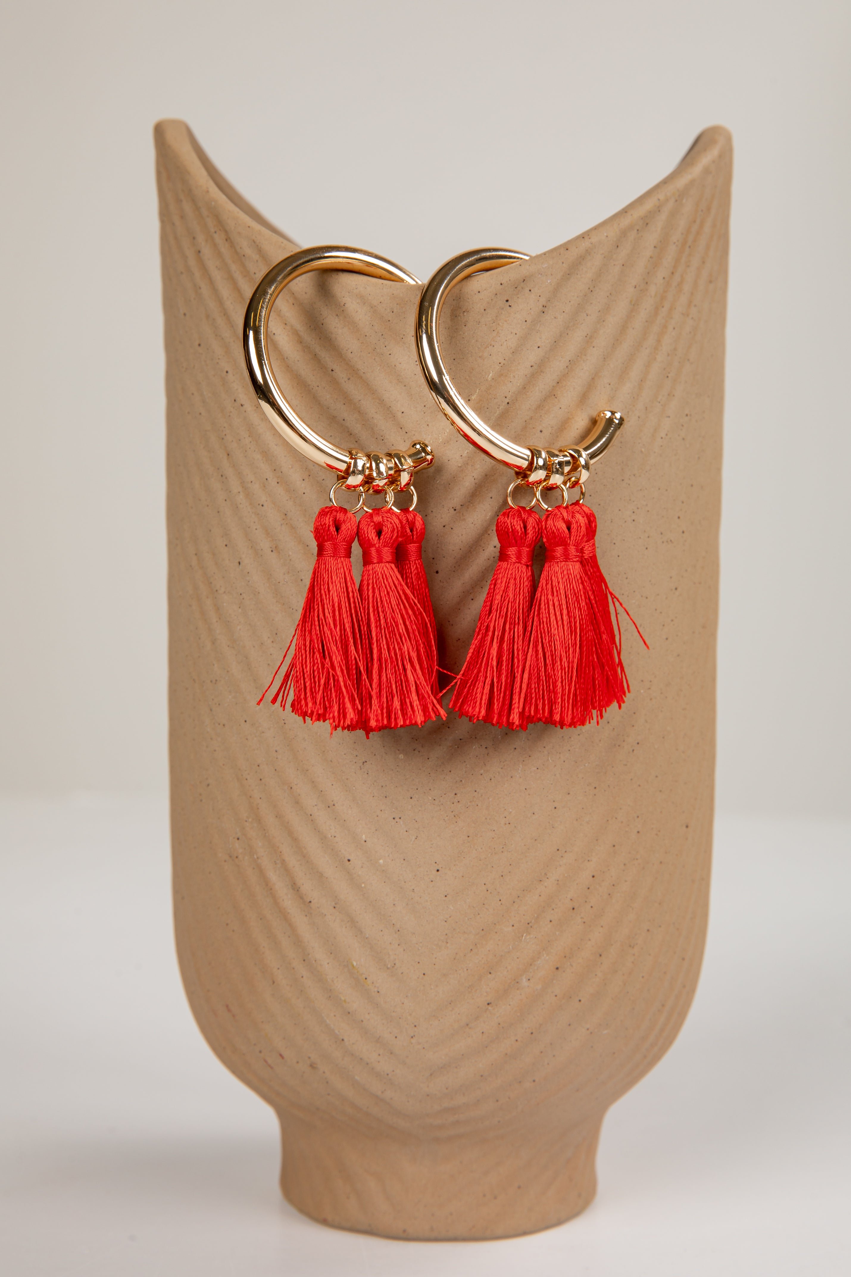 Gold hoop earrings with red tassels on a beige stand.