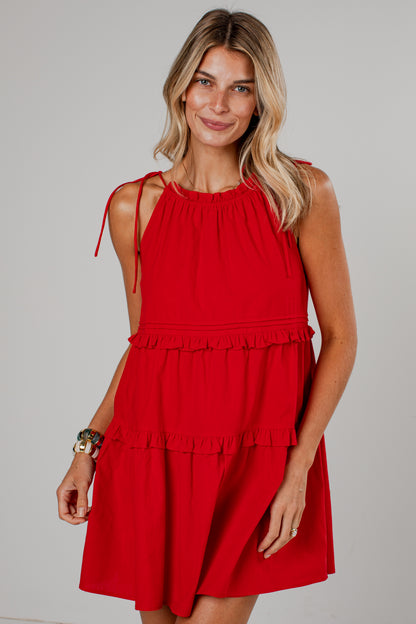 A woman modeling a bright red sleeveless tiered dress with ruffle details and tie straps on the shoulders, photographed against a neutral light gray studio background.