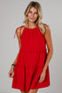A woman modeling a bright red sleeveless tiered dress with ruffle details and tie straps on the shoulders, photographed against a neutral light gray studio background.