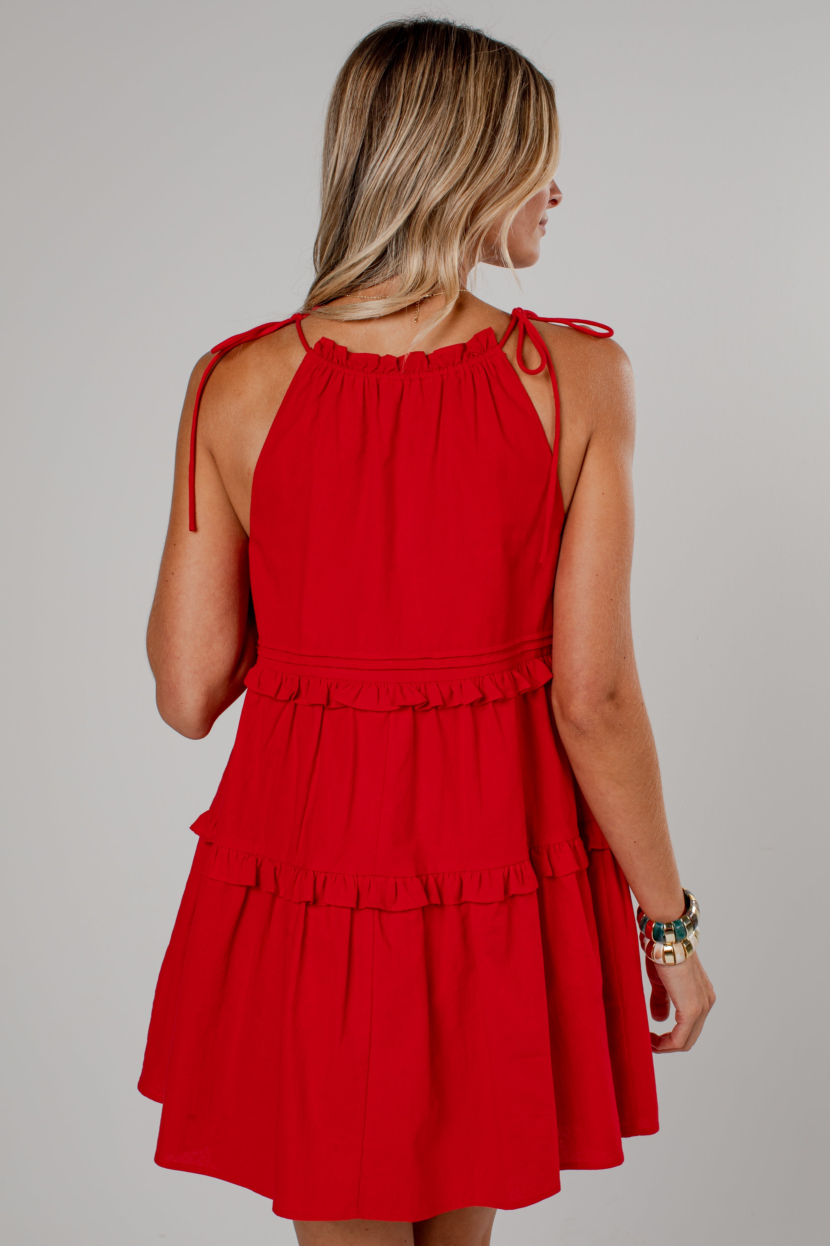A woman shown from the back wearing a bright red sleeveless tiered dress with tie straps at the shoulders, photographed against a plain light gray studio background.