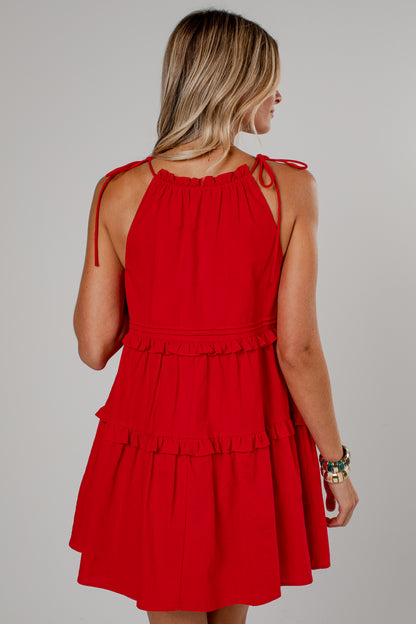 A woman shown from the back wearing a bright red sleeveless tiered dress with tie straps at the shoulders, photographed against a plain light gray studio background.