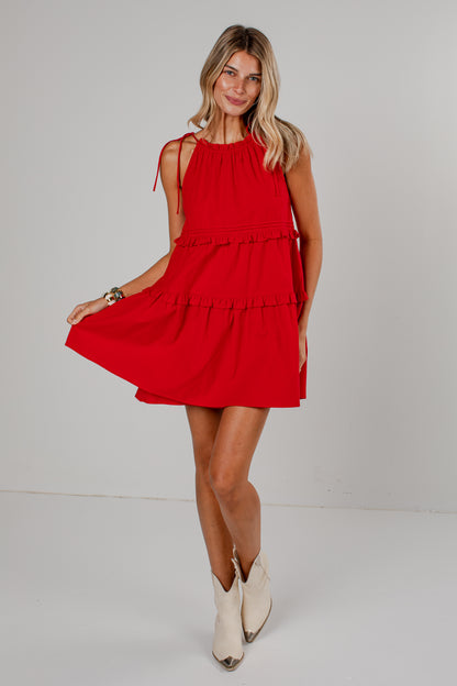 A woman modeling a bright red sleeveless tiered mini dress with tie shoulder straps, paired with cream-colored cowboy boots, against a plain light gray studio background.