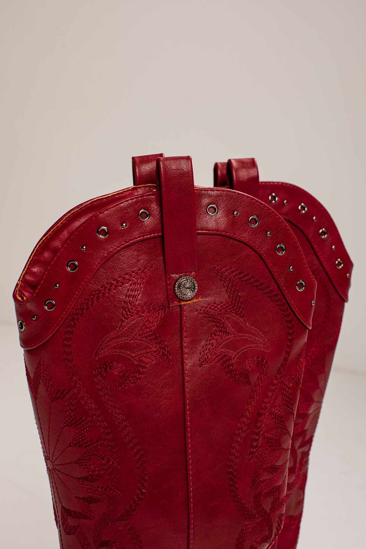 A close-up of a red leather cowboy boot showing the upper shaft with floral tooling, silver studs, and a central silver concho on a light gray background.