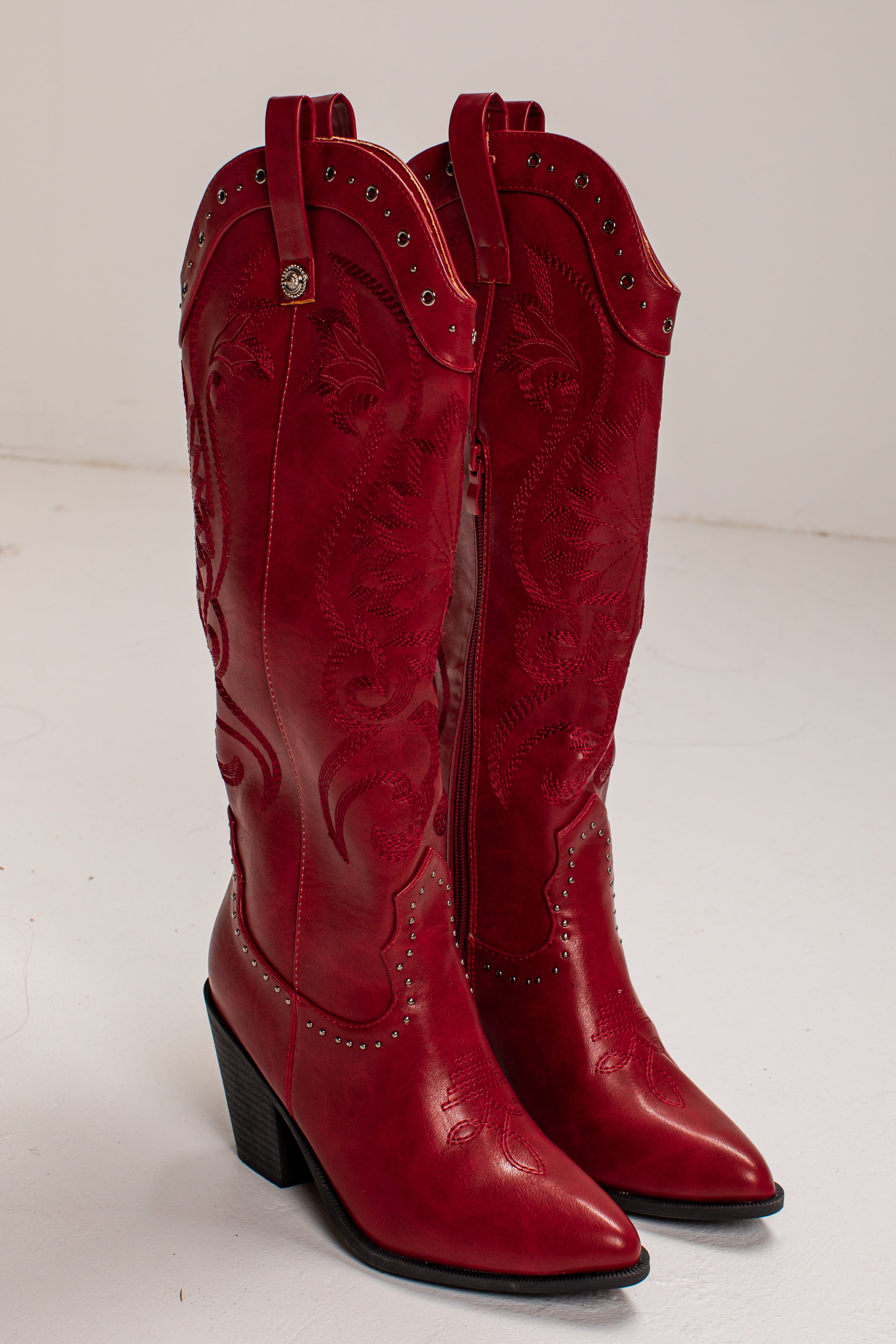 A pair of red leather cowboy boots with western embroidery, studded details, and low block heels, photographed on a light gray studio background.