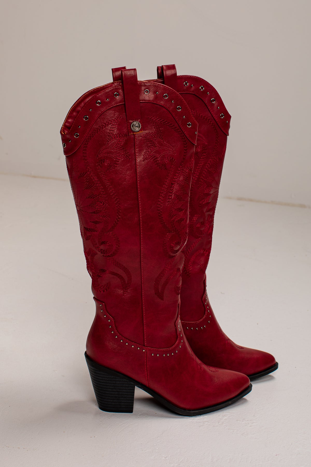 A pair of red leather cowboy boots with embossed western stitching, silver-tone studs, and black block heels, photographed against a plain off-white background.
