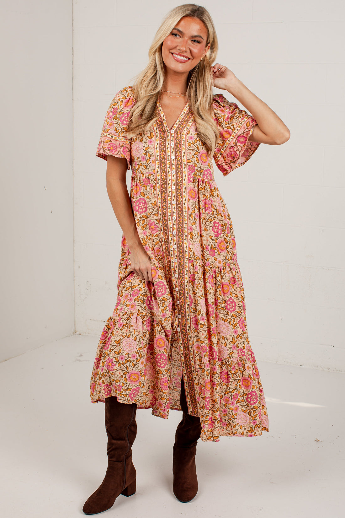 A woman modeling a short-sleeve bohemian-style midi dress with a pink, orange, and beige floral pattern, paired with brown boots, in a white studio setting.