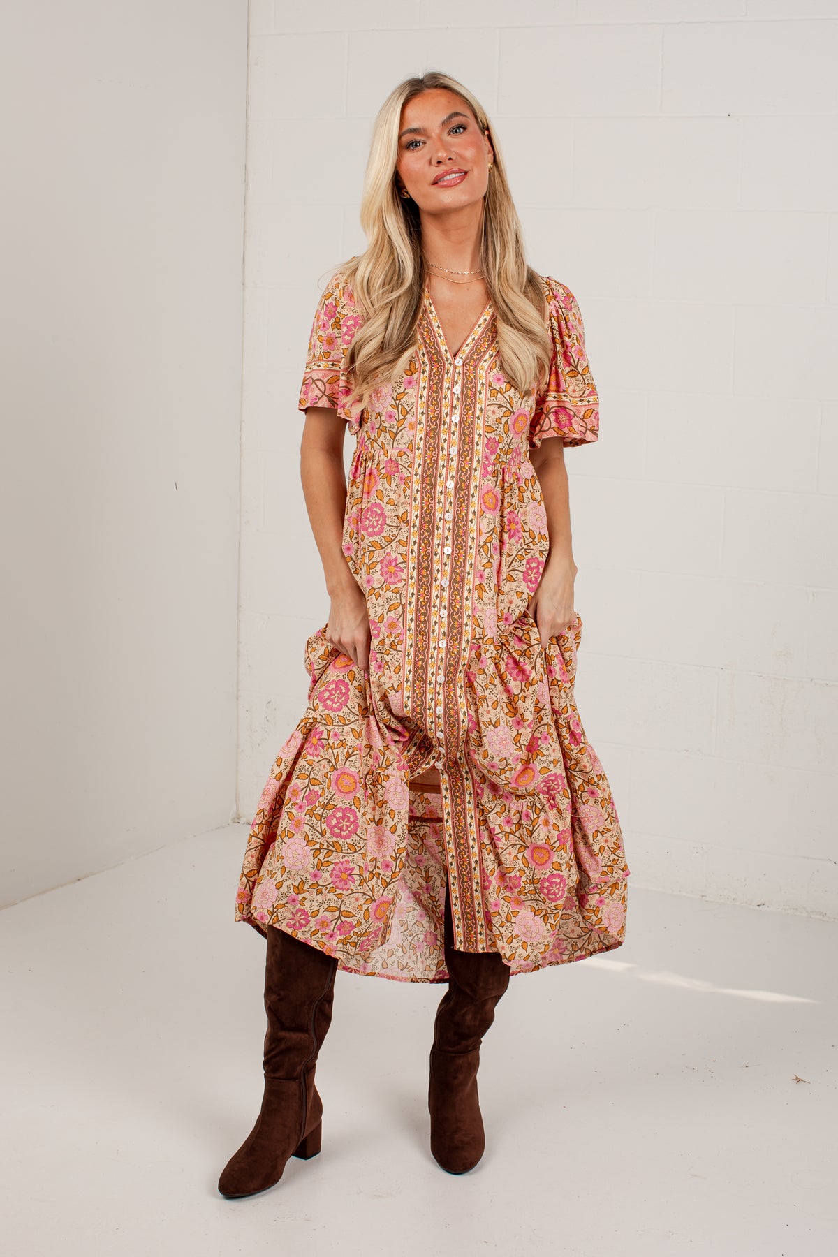 A woman modeling a short-sleeve bohemian-style midi dress with a pink, orange, and beige floral print, paired with brown knee-high boots, standing against a white studio wall.