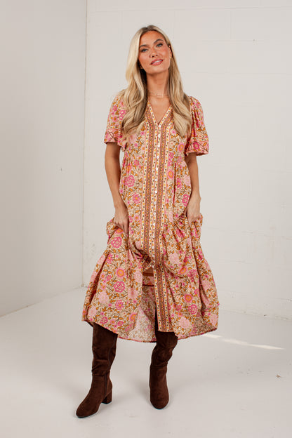 A woman modeling a short-sleeve bohemian-style midi dress with a pink, orange, and beige floral print, paired with brown knee-high boots, standing against a white studio wall.