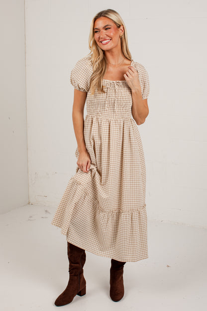 A woman modeling a beige and white gingham midi dress with puff sleeves, a smocked bodice, and tiered ruffle hem, paired with brown suede boots, in a white studio setting.