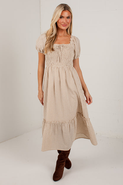 A woman modeling a beige and white gingham midi dress with puff sleeves, a smocked bodice, and a tiered skirt, paired with brown suede boots against a white studio background.