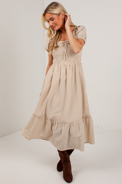 A woman modeling a beige and white gingham tiered midi dress with short puff sleeves and a smocked bodice, paired with brown suede boots, against a white studio background.
