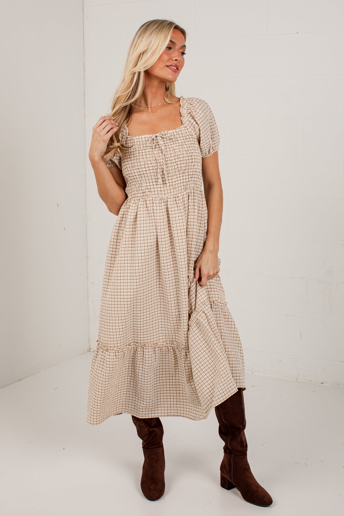 A woman modeling a beige and white gingham tiered midi dress with short puff sleeves, a square neckline, and a smocked bodice, paired with brown knee-high boots against a white studio background.