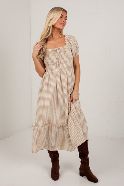 A woman modeling a beige and white gingham tiered midi dress with short puff sleeves, a square neckline, and a smocked bodice, paired with brown knee-high boots against a white studio background.