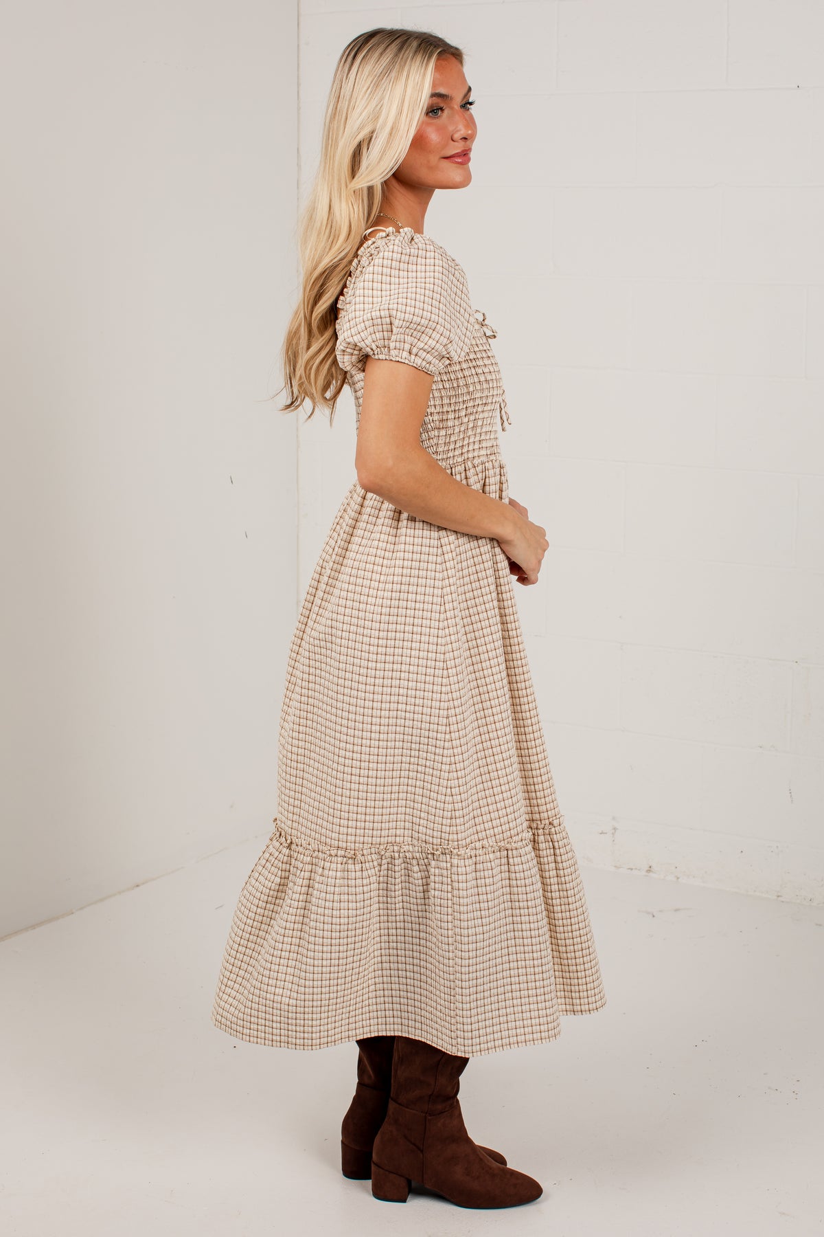 A woman modeling a beige and white gingham tiered midi dress with puff sleeves, shown from a side-back angle against a white studio background.