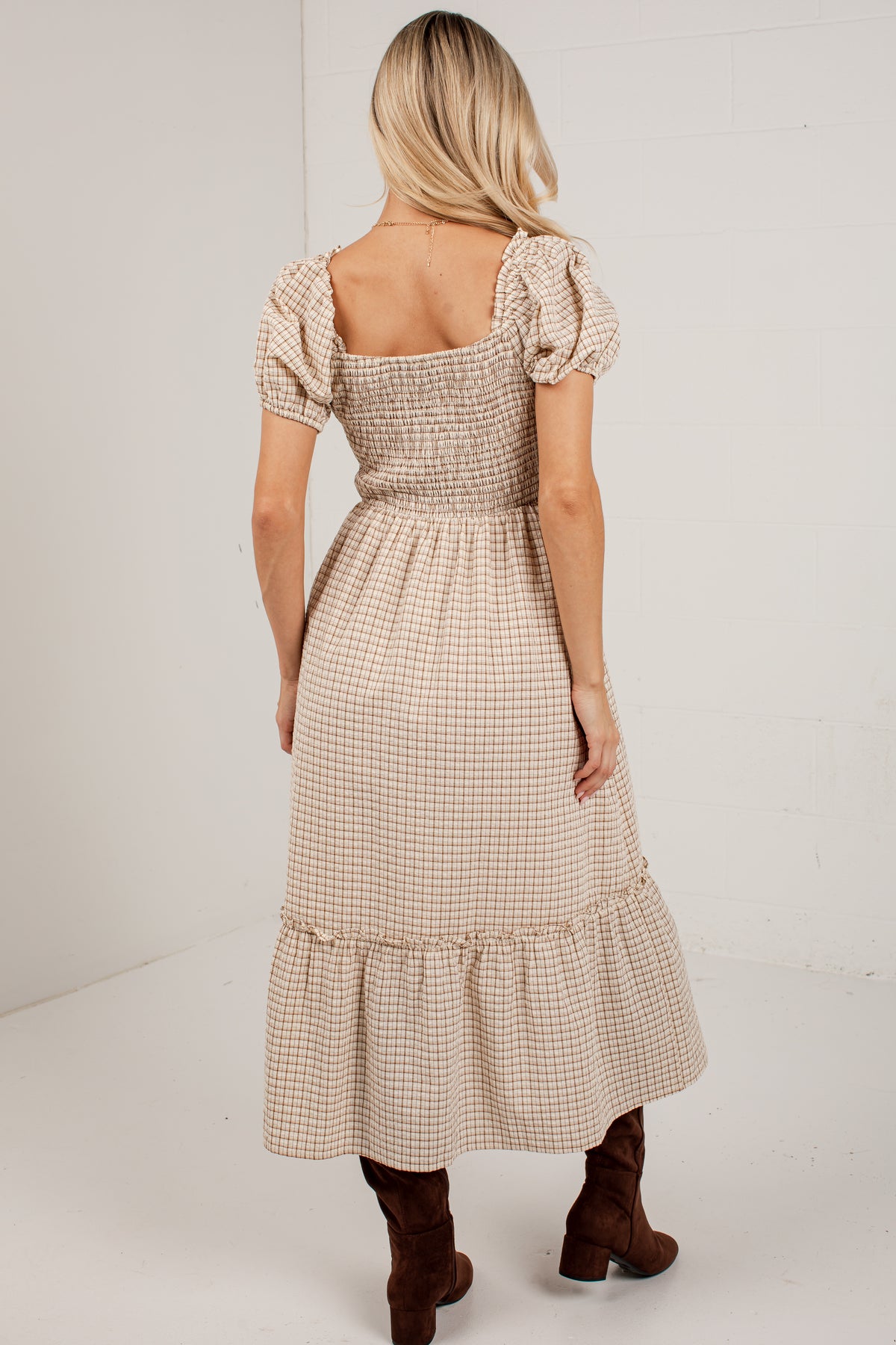 A woman viewed from the back wearing a beige and white gingham midi dress with puff sleeves and a smocked bodice, paired with brown ankle boots, against a white studio background.