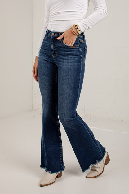 A model wearing dark blue high-waisted flare jeans with a frayed hem, paired with a white long-sleeve top and white western-style boots, standing against a plain off-white wall.