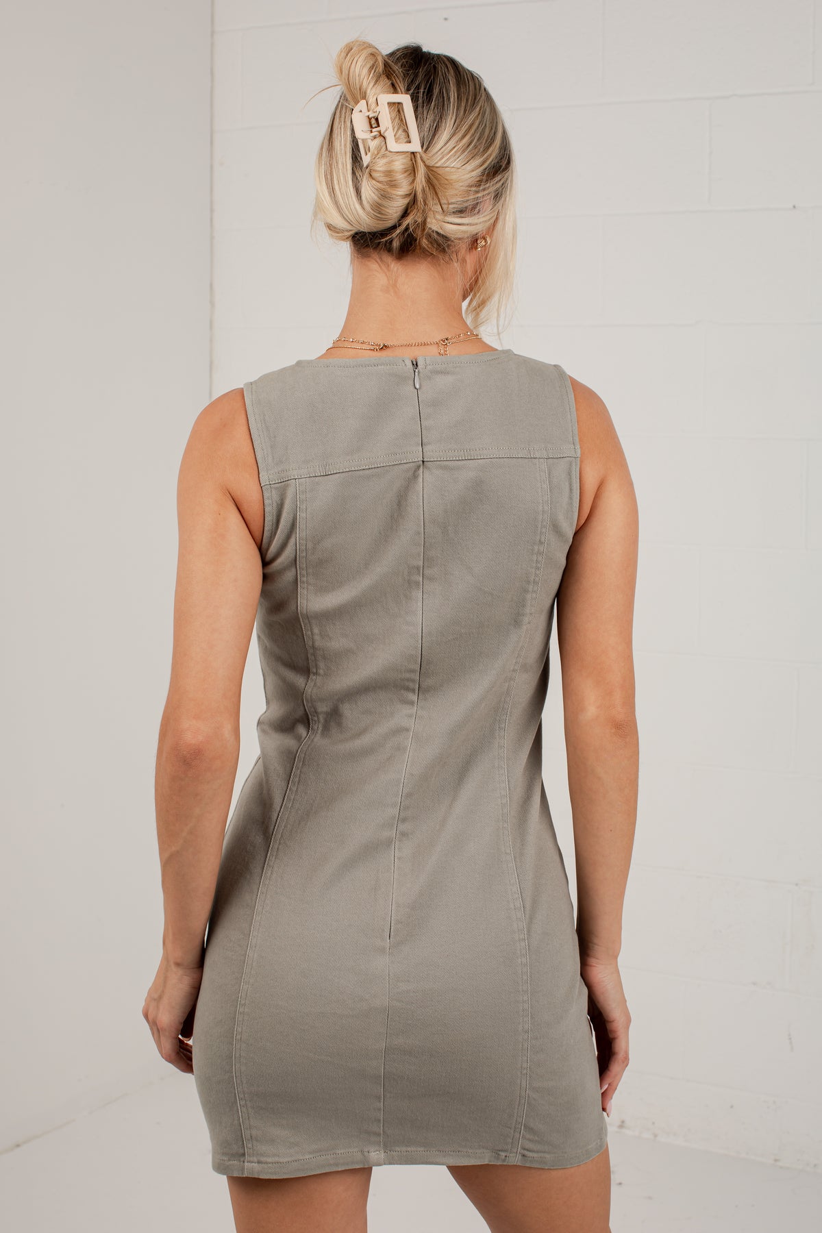 A woman viewed from behind wearing a sleeveless taupe-gray mini dress with a structured back design, set against a white brick wall.