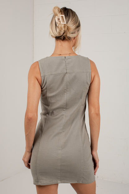 A woman viewed from behind wearing a sleeveless taupe-gray mini dress with a structured back design, set against a white brick wall.