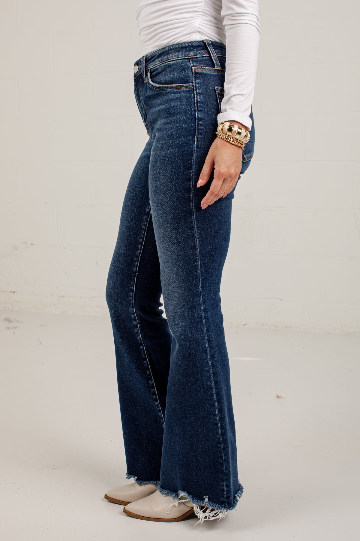 A person wearing dark blue high-waisted flare jeans with a raw frayed hem, styled with a white long-sleeve top and beige cowboy boots against a white studio background.