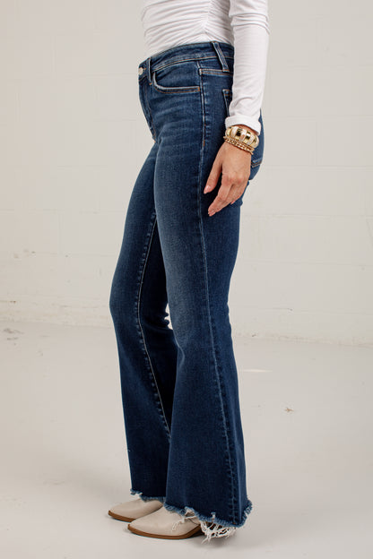 A person wearing dark blue high-waisted flare jeans with a raw frayed hem, styled with a white long-sleeve top and beige cowboy boots against a white studio background.