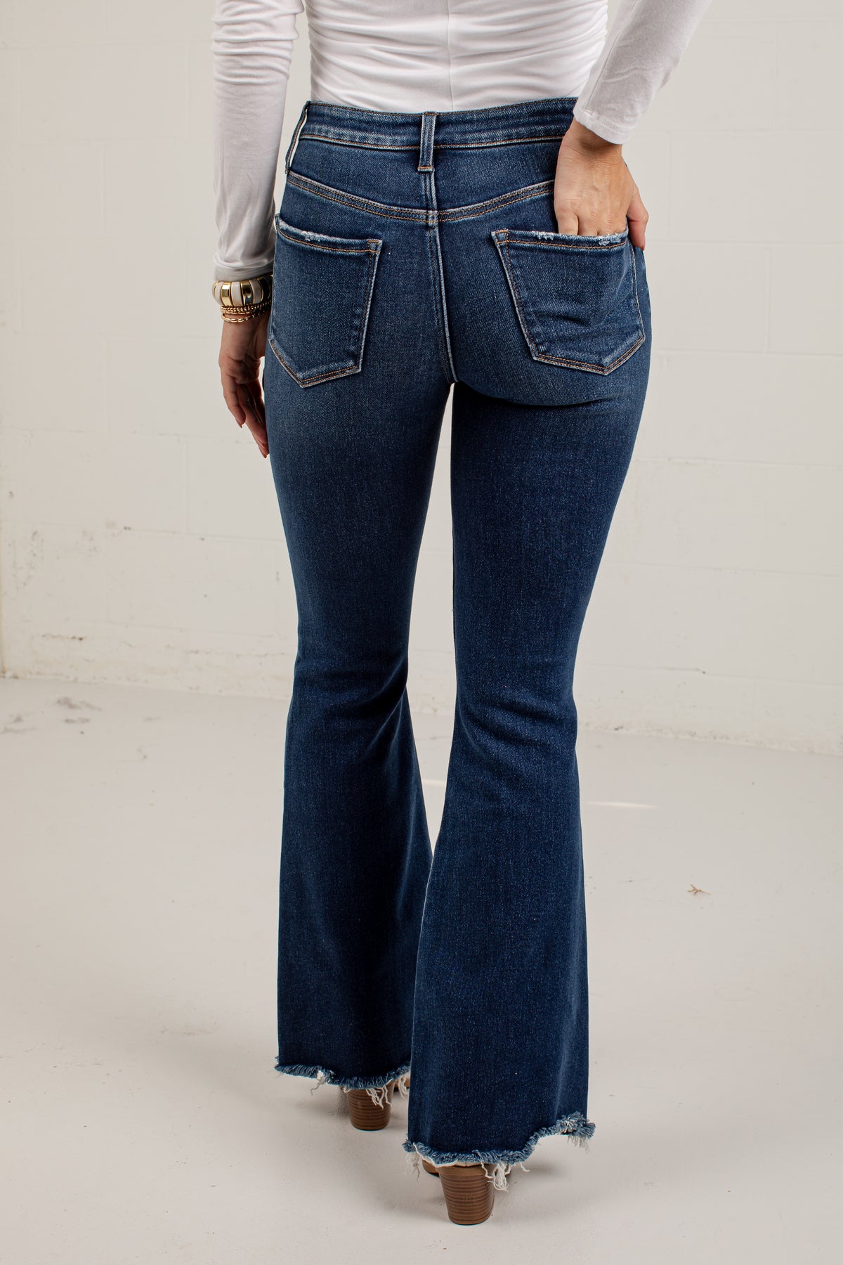 A person shown from the back wearing dark blue flare jeans with a frayed hem, paired with a white long-sleeve top and brown heeled shoes, standing against a plain off-white wall.