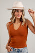 A woman wearing a rust-orange ribbed V-neck tank top, blue jeans, and a cream wide-brim fedora hat, posing against a white paneled wall.