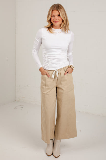 A woman modeling a white long-sleeve ruched top and beige wide-leg cropped pants with a drawstring waist, standing against a plain off-white studio wall.