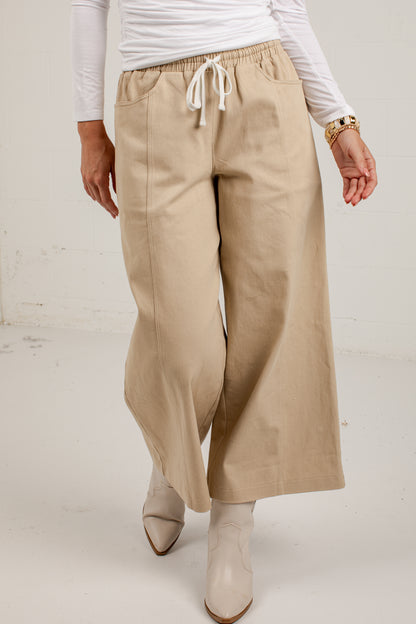 A person wearing wide-leg beige pants with a white drawstring waist, paired with a white long-sleeve top and light beige ankle boots, shown from the chest down against a plain off-white studio background.