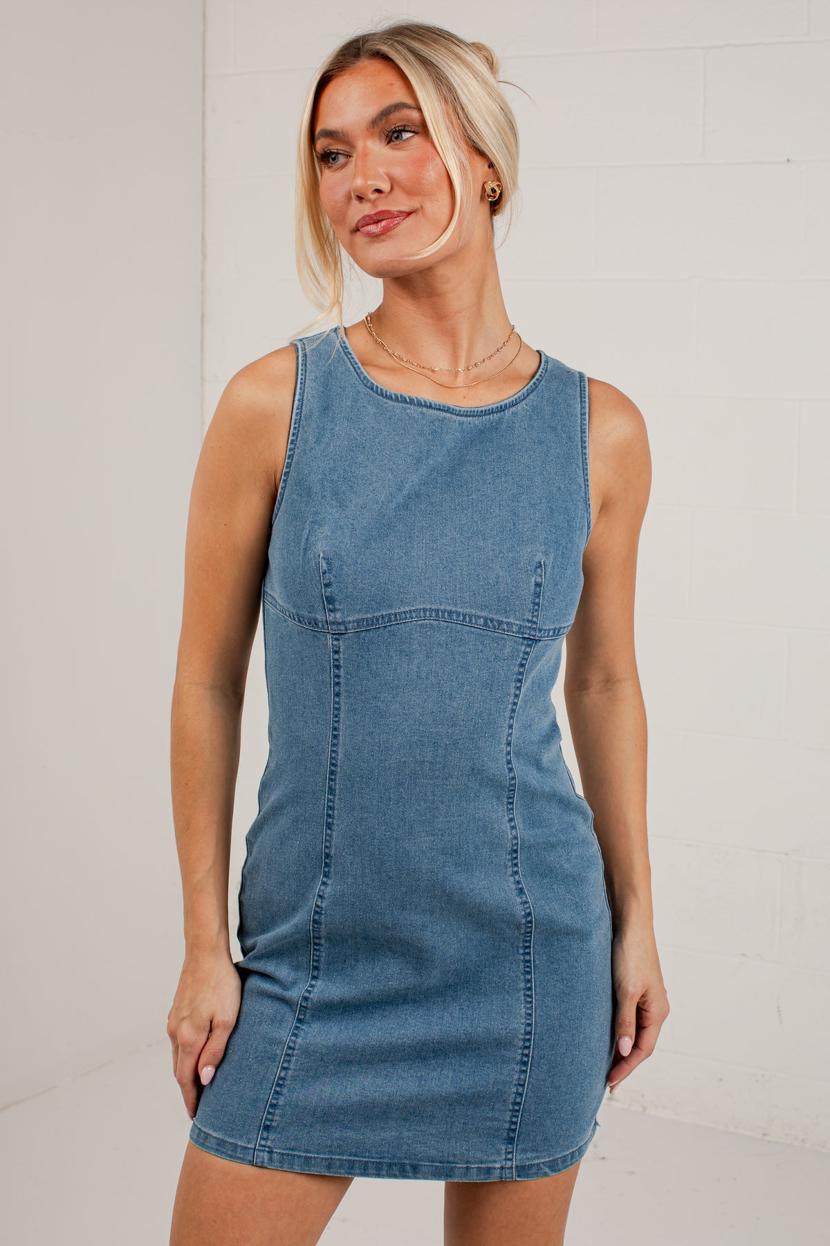 A woman modeling a sleeveless blue denim mini dress against a white brick wall background.