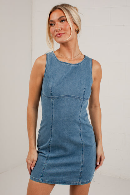 A woman modeling a sleeveless blue denim mini dress against a white brick wall background.