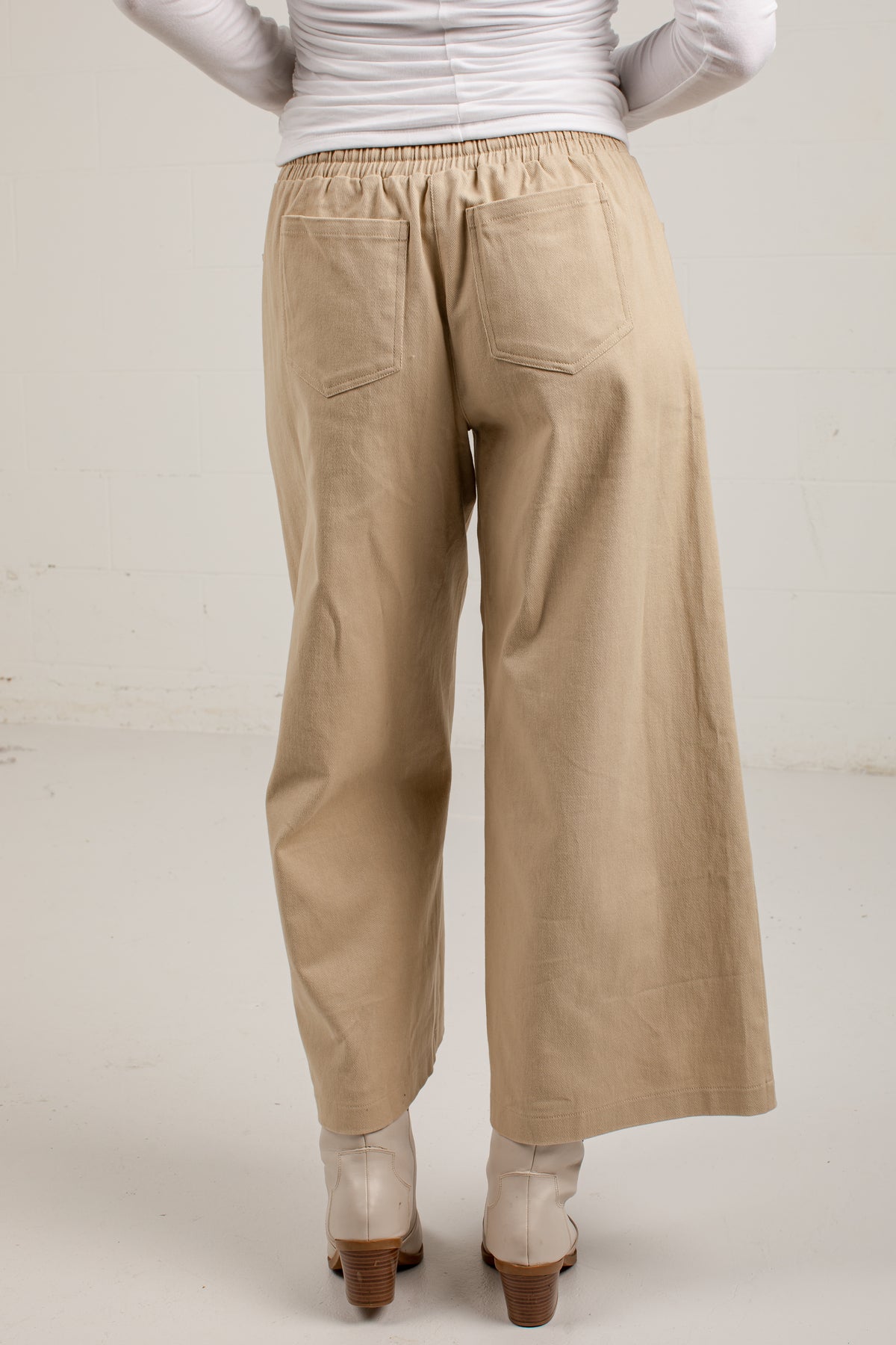 A person shown from the back wearing loose, wide-leg beige pants with an elastic waistband and back pockets, paired with a white ribbed long-sleeve top and light-colored heeled boots against a plain off-white studio background.