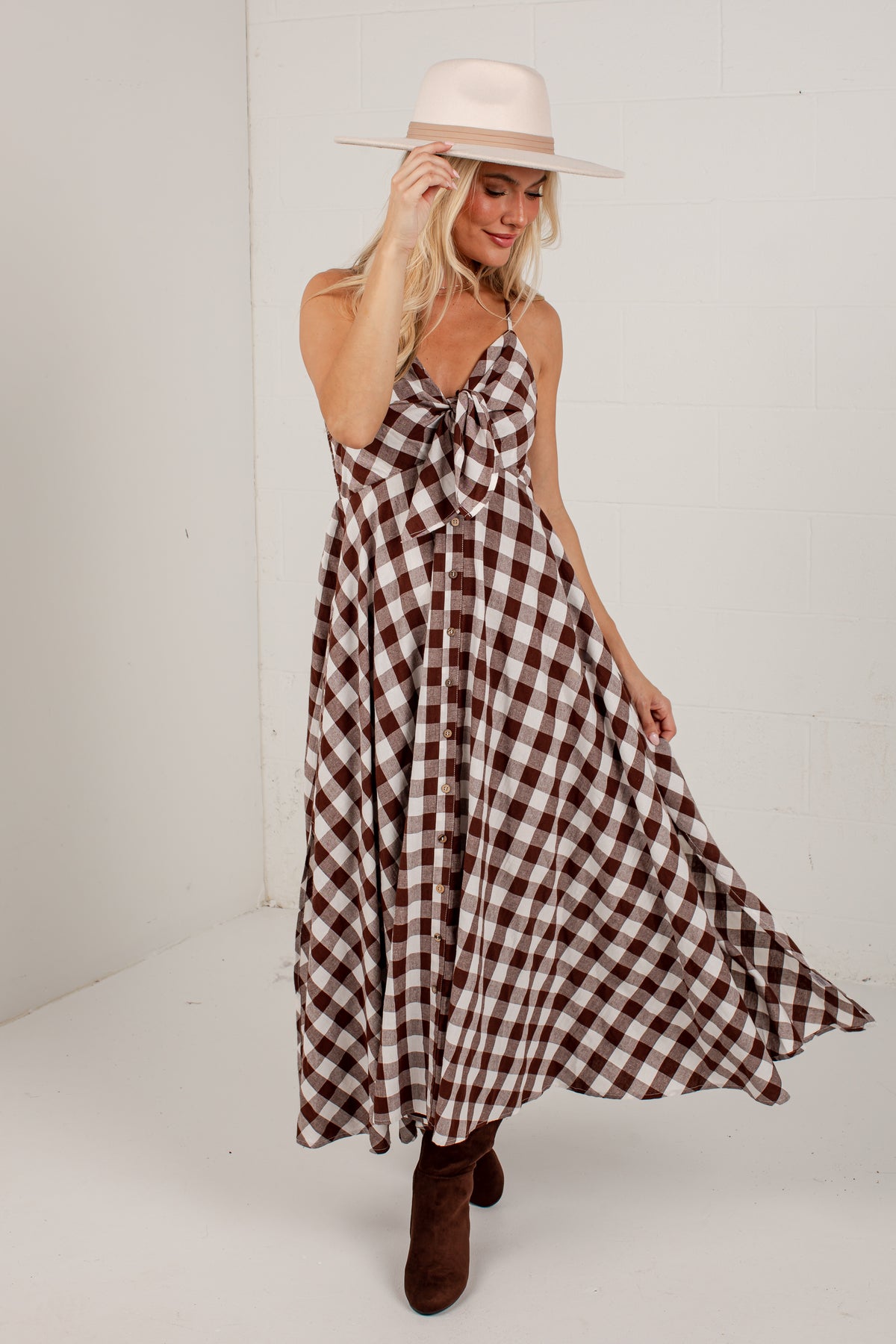 A woman modeling a brown and white gingham maxi dress with a tie-neck detail, paired with a cream wide-brim hat and brown ankle boots, set against a plain white studio background.
