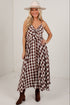 A woman modeling a brown and beige gingham maxi dress with spaghetti straps and a front tie detail, paired with a beige wide-brim hat and brown ankle boots, in a white studio setting.
