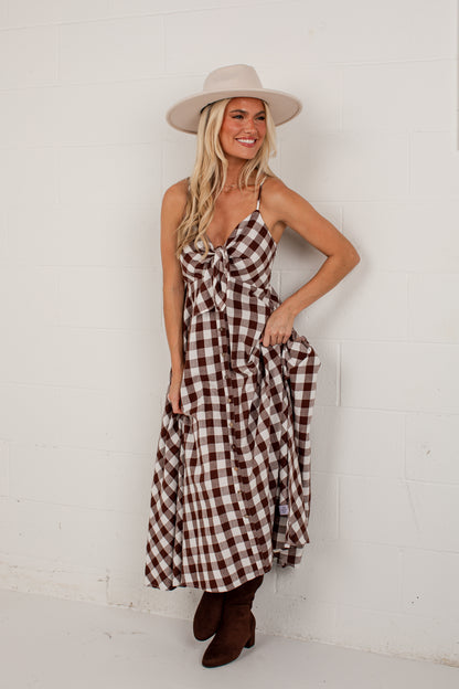 A woman modeling a brown and white gingham midi dress with a knotted sweetheart neckline, wearing a beige wide-brim hat and brown boots, posing against a white brick wall.