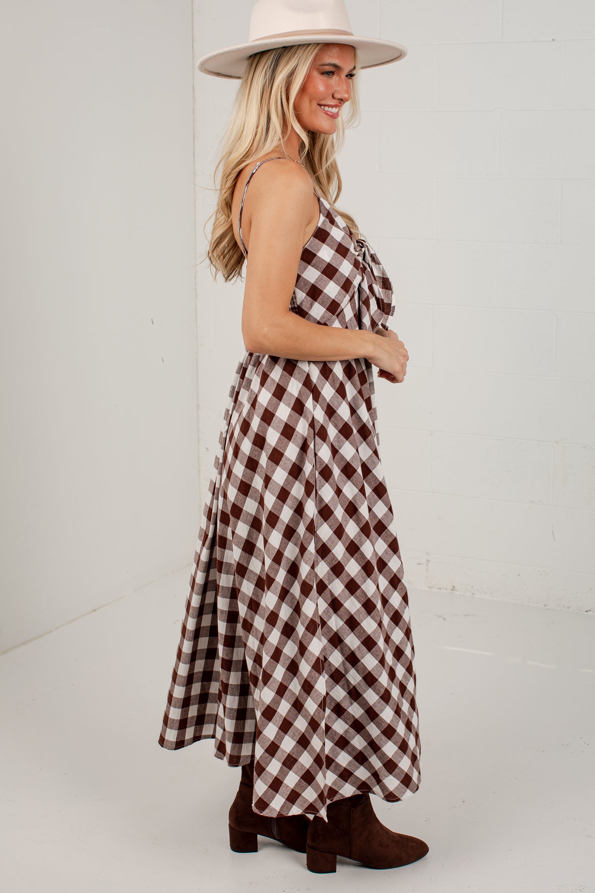 A woman wearing a brown and white gingham maxi dress with a tie-shoulder design, paired with a cream wide-brim hat and brown ankle boots, standing against a white studio wall.