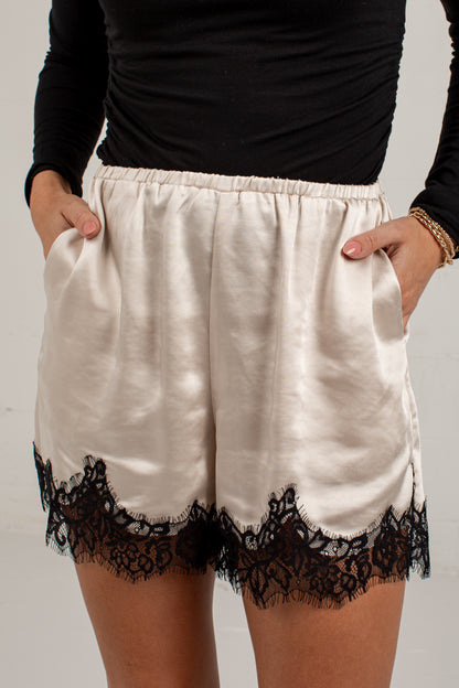 A person wearing cream satin shorts with black lace trim at the hem, paired with a black long-sleeve top. The photo is cropped from the chest down against a plain light background.