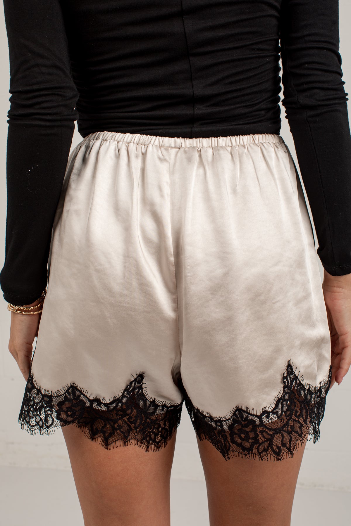 A person wearing satin-like shorts in a warm off-white or cream tone with black lace trim at the hem, paired with a black long-sleeve top. The photo is cropped from the chest to the upper thighs against a plain light background.