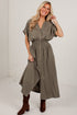A woman modeling a short-sleeve, button-front midi dress in a muted olive green color, paired with brown suede boots, standing against a white studio wall.