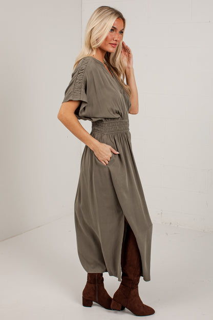 A woman modeling a loose, short-sleeve olive green maxi dress with a smocked waist and brown suede ankle boots, standing against a plain white studio wall.