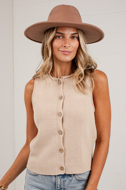 A woman wearing a beige sleeveless knit button-front vest, a brown wide-brim fedora hat, and light-wash blue jeans, standing against a white brick wall.