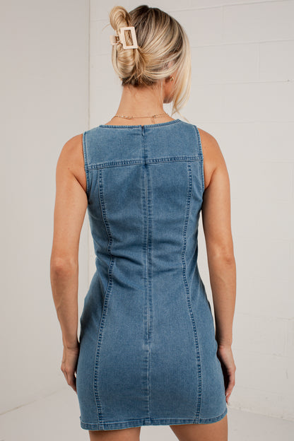 A woman viewed from behind wearing a sleeveless blue denim dress, standing against a light-colored brick wall.