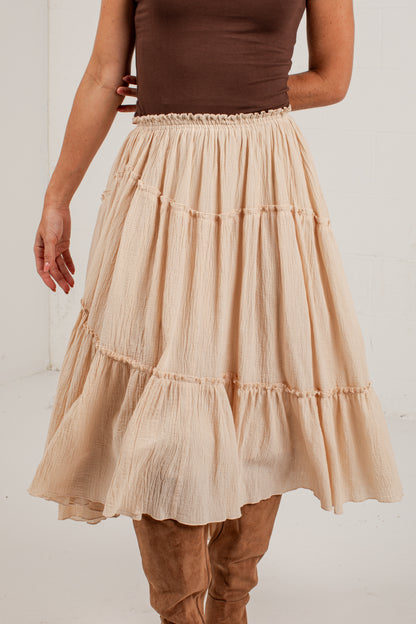 A beige tiered gauze midi skirt worn by a model, paired with a brown top and brown suede boots, shown from the chest down against a white studio background.