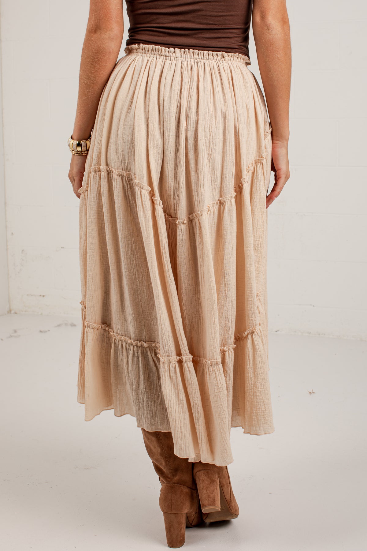 A person shown from the back wearing a long tiered midi skirt in a light warm beige or blush-tan tone, paired with a dark brown top and brown heeled boots, against a plain off-white wall.