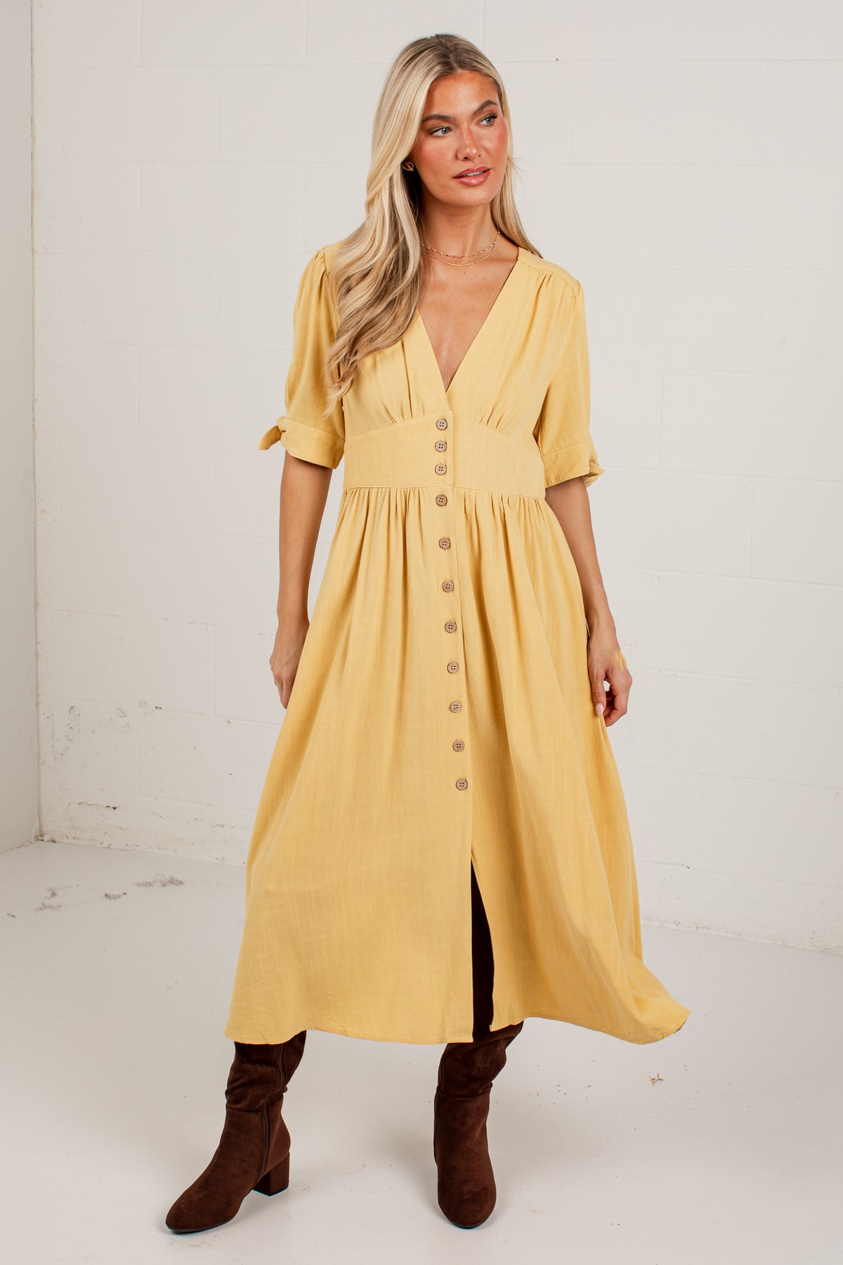 A woman modeling a mustard yellow midi dress with short sleeves, a V-neckline, and a button-down front, paired with brown knee-high boots against a white studio background.