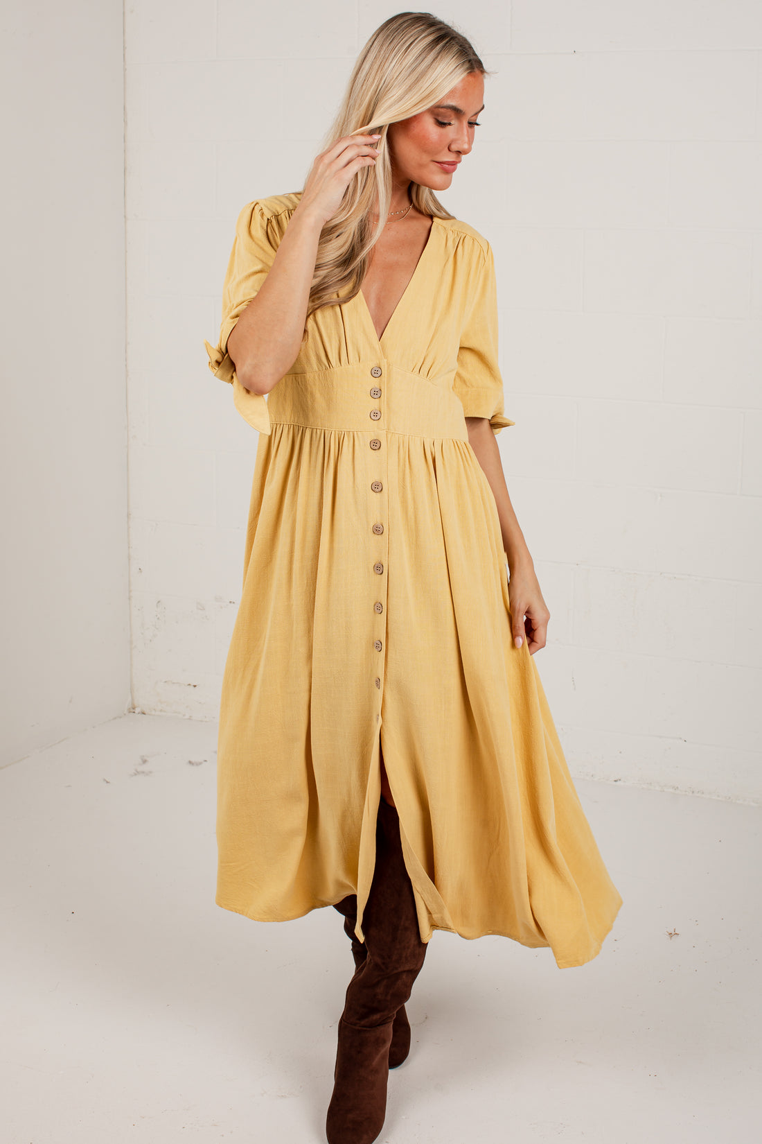A woman modeling a mustard yellow midi dress with puff sleeves, a V-neckline, and a button-down front, paired with brown boots against a plain white studio background.
