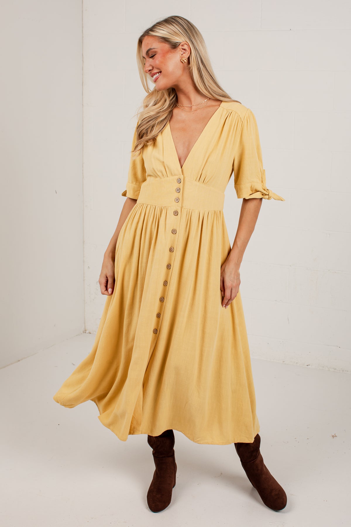 A woman modeling a yellow midi dress with a button-down front, short sleeves with tie details, and a gathered waist, paired with brown ankle boots in a bright studio setting.